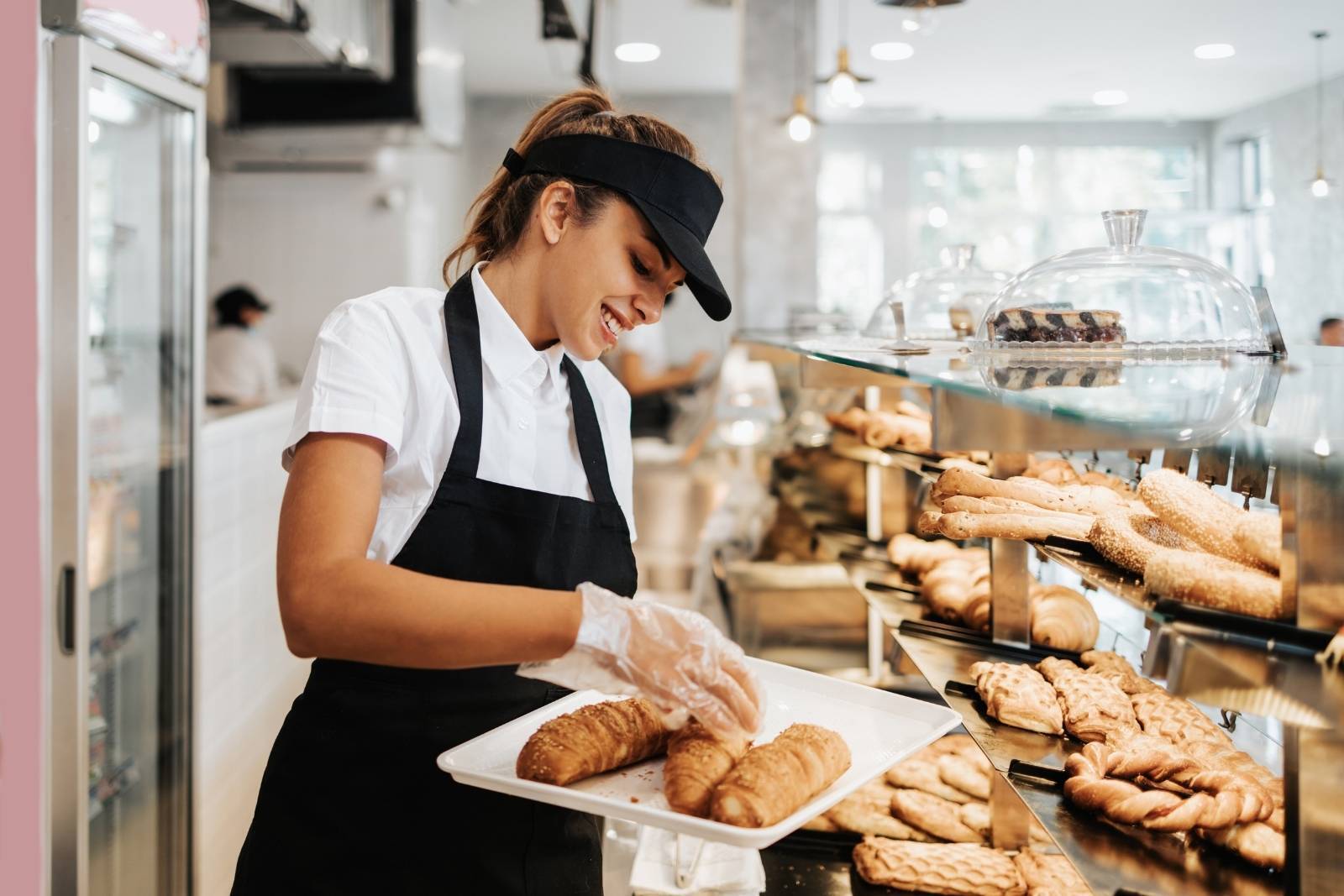 A foodservice professional in a hat and apron holds a tray with loaves of bread behind the counter of a bakery.